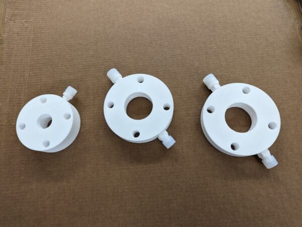 Three PTFE/HDPE flanges with connecting nozzles laid out in a row on a cardboard background.